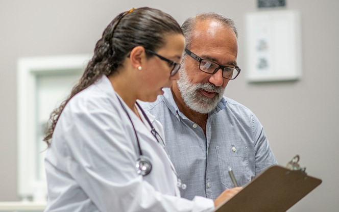 A Female Doctor Sits Next To An Elderly Male Patient Reviewing The Patient Chart Together